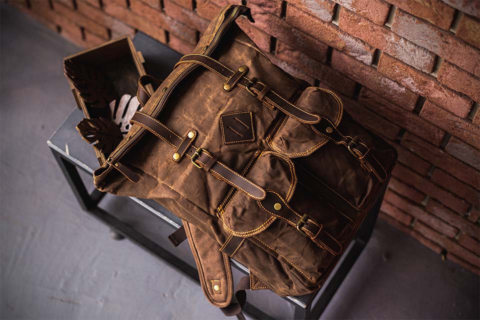 Brown waxed canvas backpack with leather straps on a stool by a brick wall