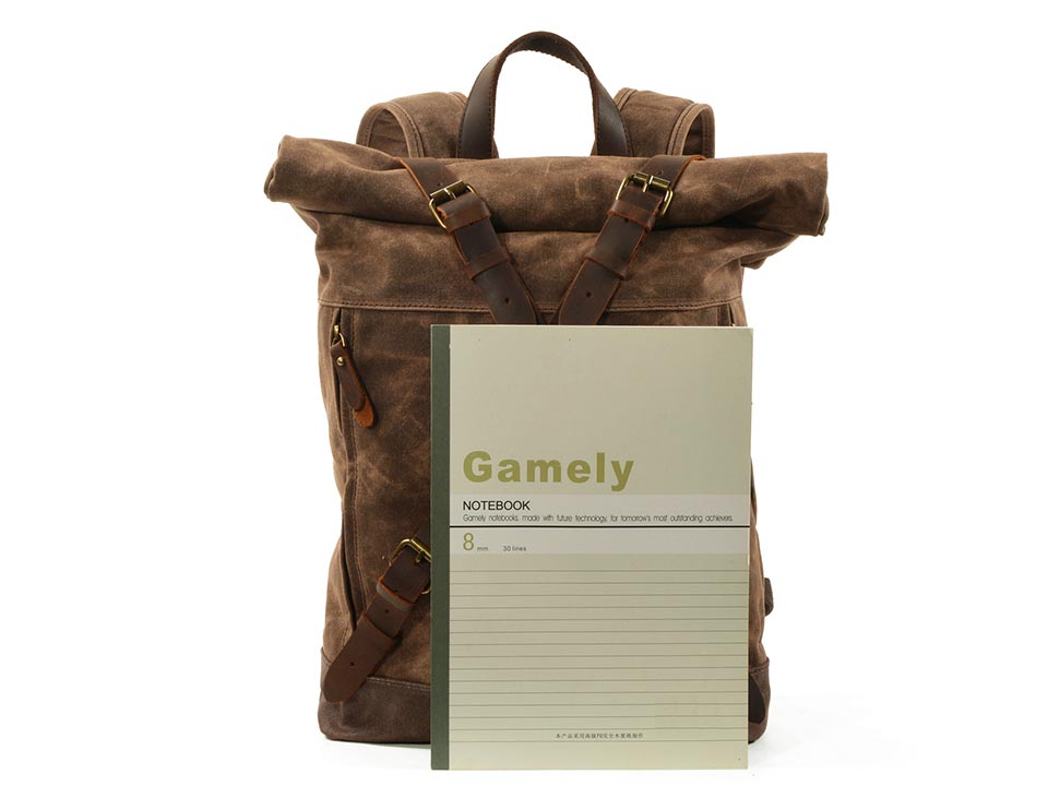 Coffee waxed canvas roll top backpack beside a paper notebook for size comparison