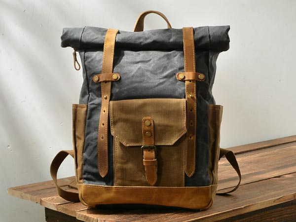 Waxed canvas roll top backpack with leather buckle straps standing on a wooden surface