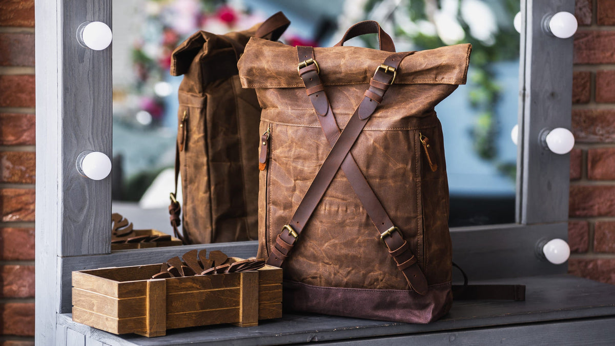 Two brown waxed canvas roll-top backpacks on a table by a lit mirror