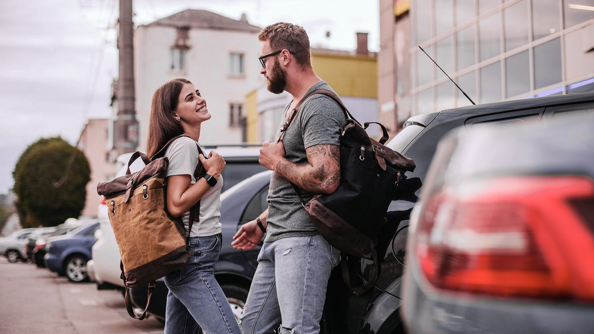 A man and a woman talking on a city street, the woman wearing a waxed canvas roll top backpack
