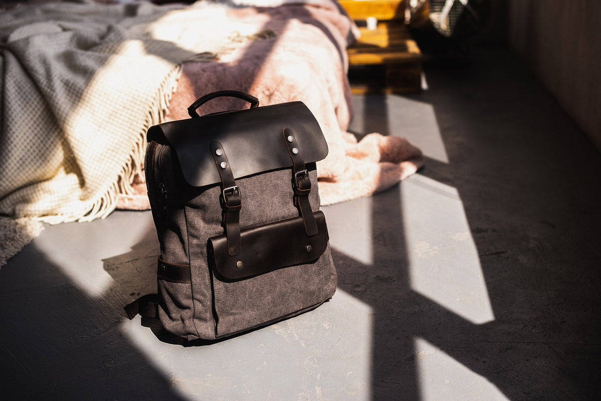 Gray canvas backpack with a black leather flap standing on a sunlit floor