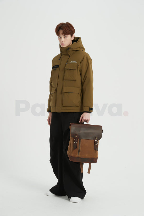 Man standing with a khaki waxed canvas backpack with a leather flap, holding its top handle in his left hand