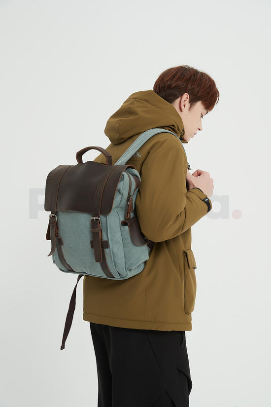 Side view of a man facing right and looking down while wearing a blue waxed canvas backpack with a leather flap