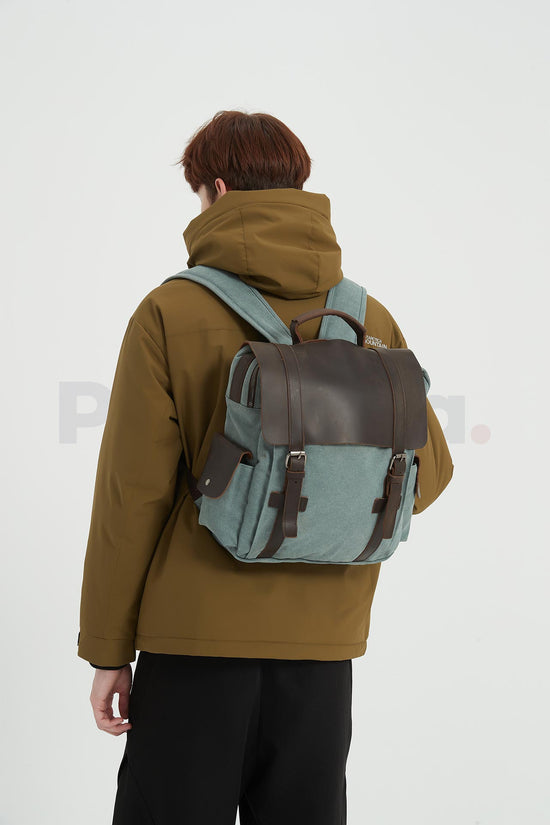 Rear three-quarter view of a man wearing a blue waxed canvas backpack with a leather flap and facing left