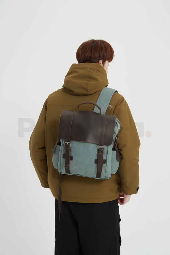 Rear three-quarter view of a man wearing a blue waxed canvas backpack with a leather flap and facing right