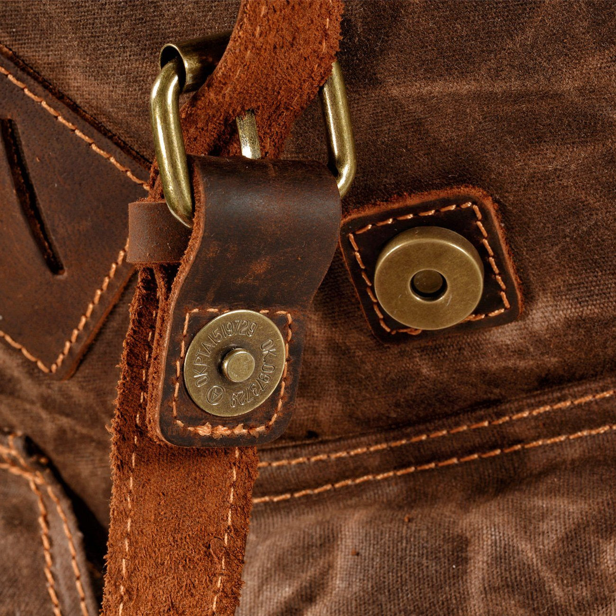 Open magnetic snap beneath a leather buckle tab on the front pocket of coffee waxed canvas backpack.